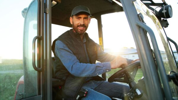 Caucasian male farmer in cap sitting in tractor with open door and smiling to camera