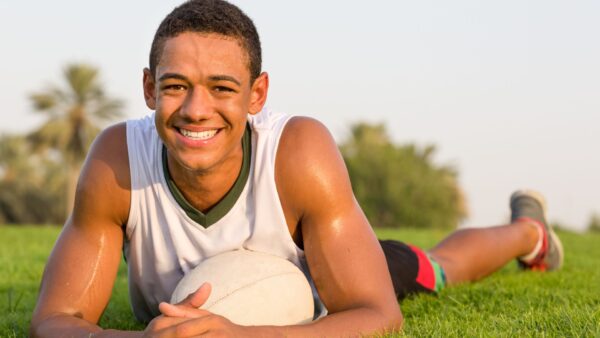 Happy fit black teen athlete resting on the grass with a rugby ball