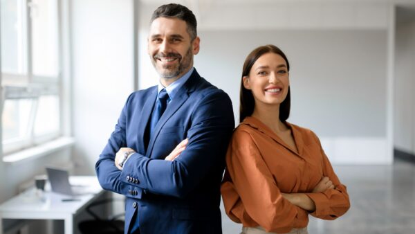 Successful businessman and businesswoman standing back to back with arms crossed and smiling at camera, office interior