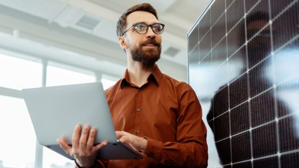Smiling man, engineer, developer holding laptop computer planning startup working with solar panel in modern office. Innovation, renewable energy concept